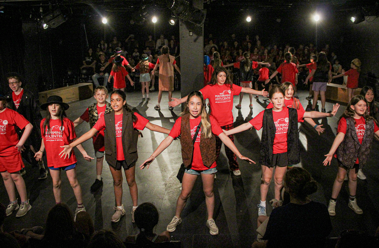 Performers on stage at Wolf Performing Arts Center, children are wearing red shirts with brown vests.